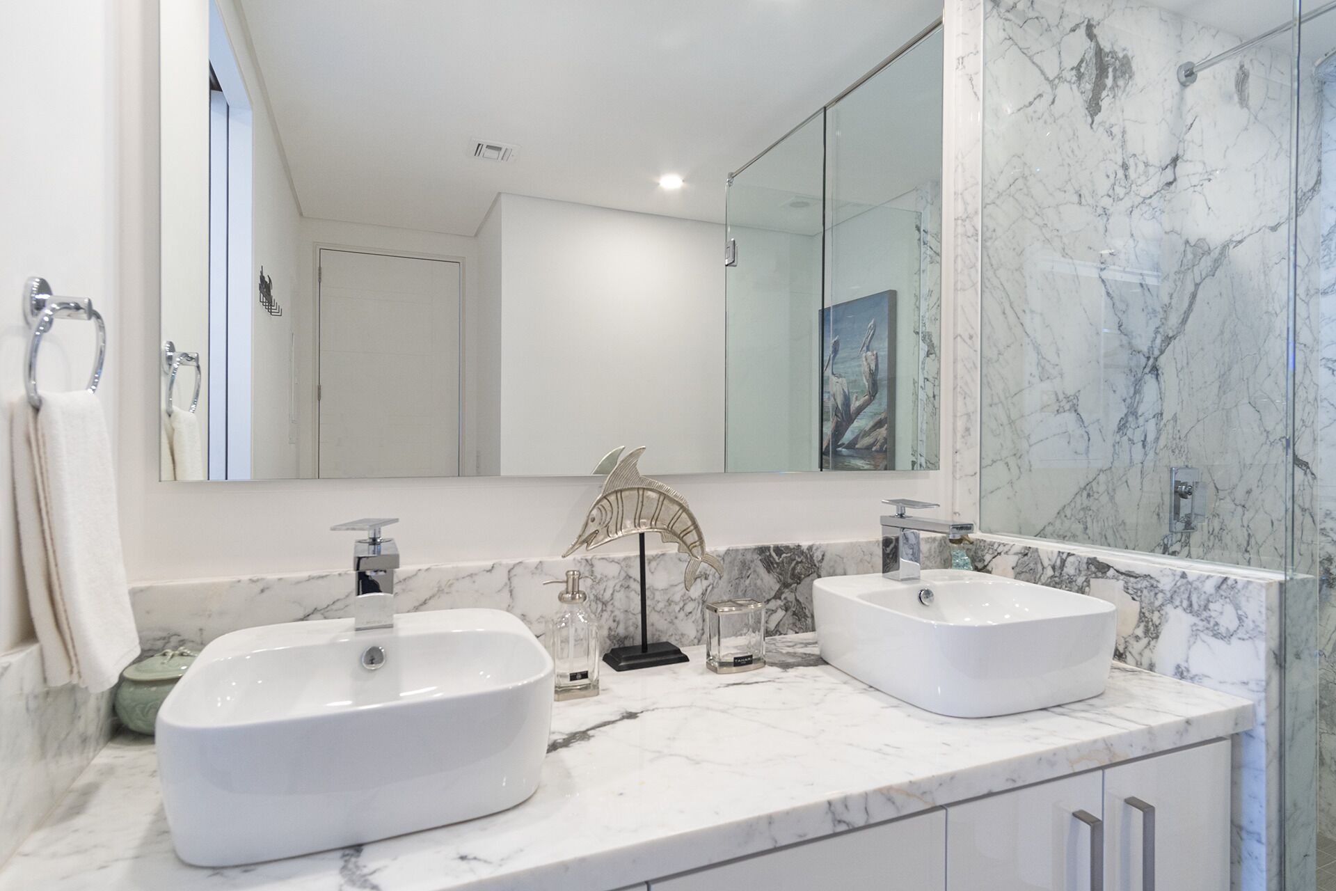 Un baño con dos lavamanos blancos, un espejo grande y una ducha con paredes de mármol.