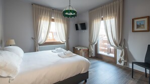 Una camera da letto con un letto, due finestre con tende, un termosifone, una TV appesa al muro e una sedia.