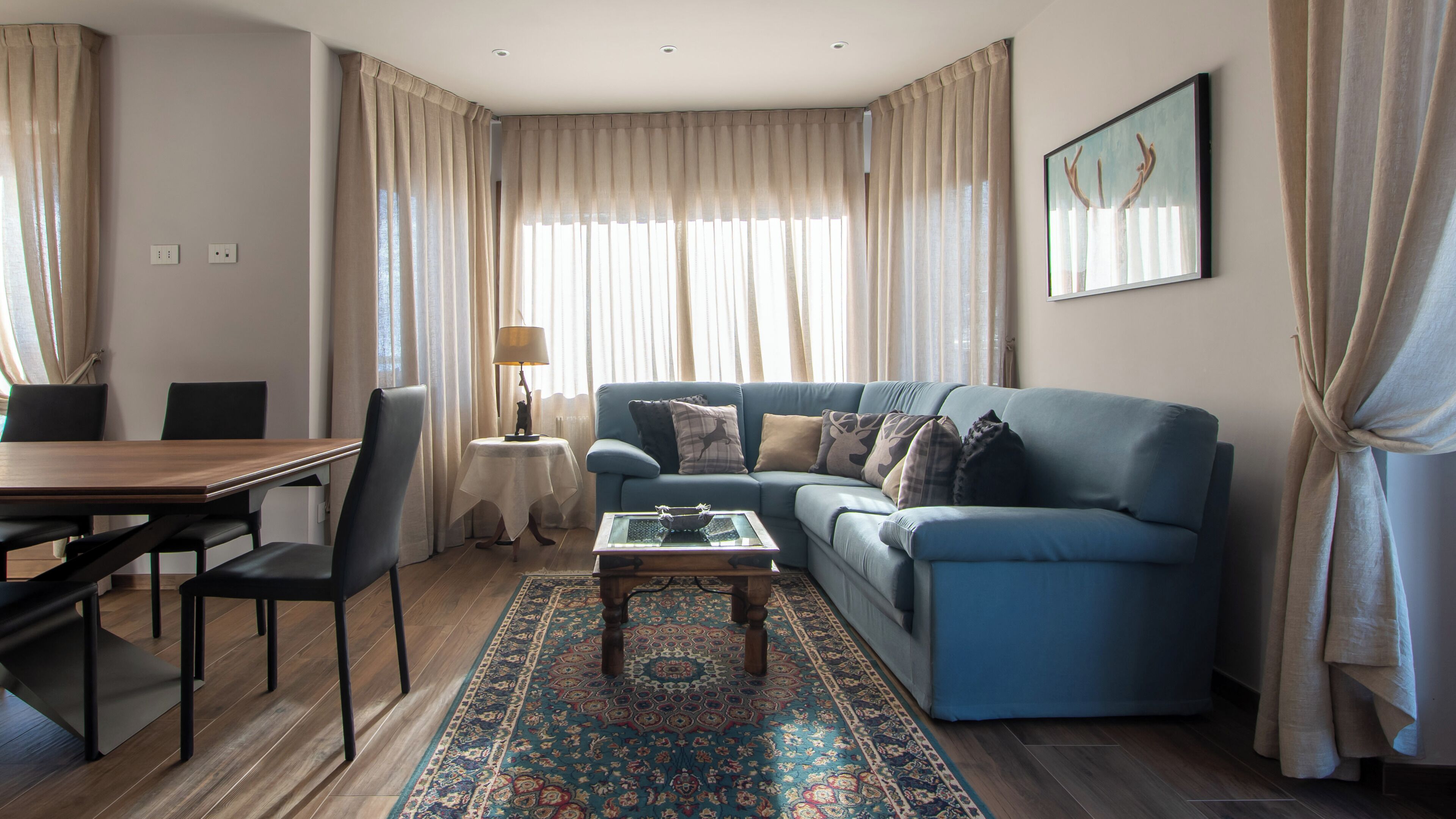 A living room with a blue sofa, a coffee table, a patterned rug, and a dining area with a wooden table and chairs.