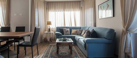 A living room with a blue sofa, a coffee table, a patterned rug, and a dining area with a wooden table and chairs.