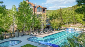 A modern building with a pool area, including a lap pool and a hot tub, surrounded by trees and a hilly landscape. - Greystone 60 Five Bedroom (Beaver Creek)