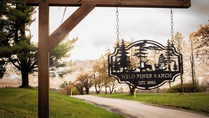 Et træskilt med et metalmærke for Wild Pines Ranch, grundlagt i 2025, hænger på en pæl ved vejen.