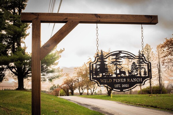 A wooden sign with a metal emblem for Wild Pines Ranch, established in 2025, hanging on a post by a road.