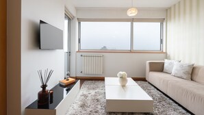 A modern living room with a sofa, a coffee table, a TV mounted on the wall, and a large window with a view of the city. - Modern Flat with Balcony & Garage by HostWise (Porto)