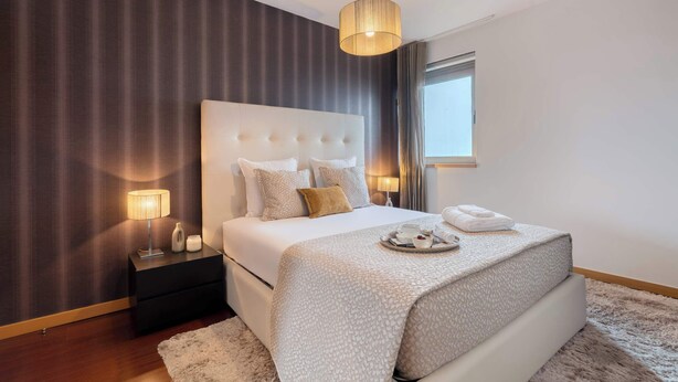 A modern bedroom with a large bed, a bedside table with a lamp, and a window with curtains. - Modern Flat with Balcony & Garage by HostWise (Porto)