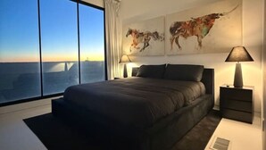 Une chambre moderne avec un grand lit, deux tables de chevet, une œuvre d’art représentant une carte du monde et une vue sur le paysage urbain, visible à travers de grandes fenêtres.