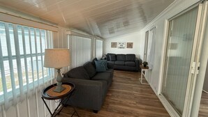 A cozy living room with a gray sofa, a small side table with a lamp, and a wooden floor. There are large windows with blinds, a sliding glass door, and a wall with a 'Relax and Unwind' sign. - Serenity House for Two! (Kissimmee)