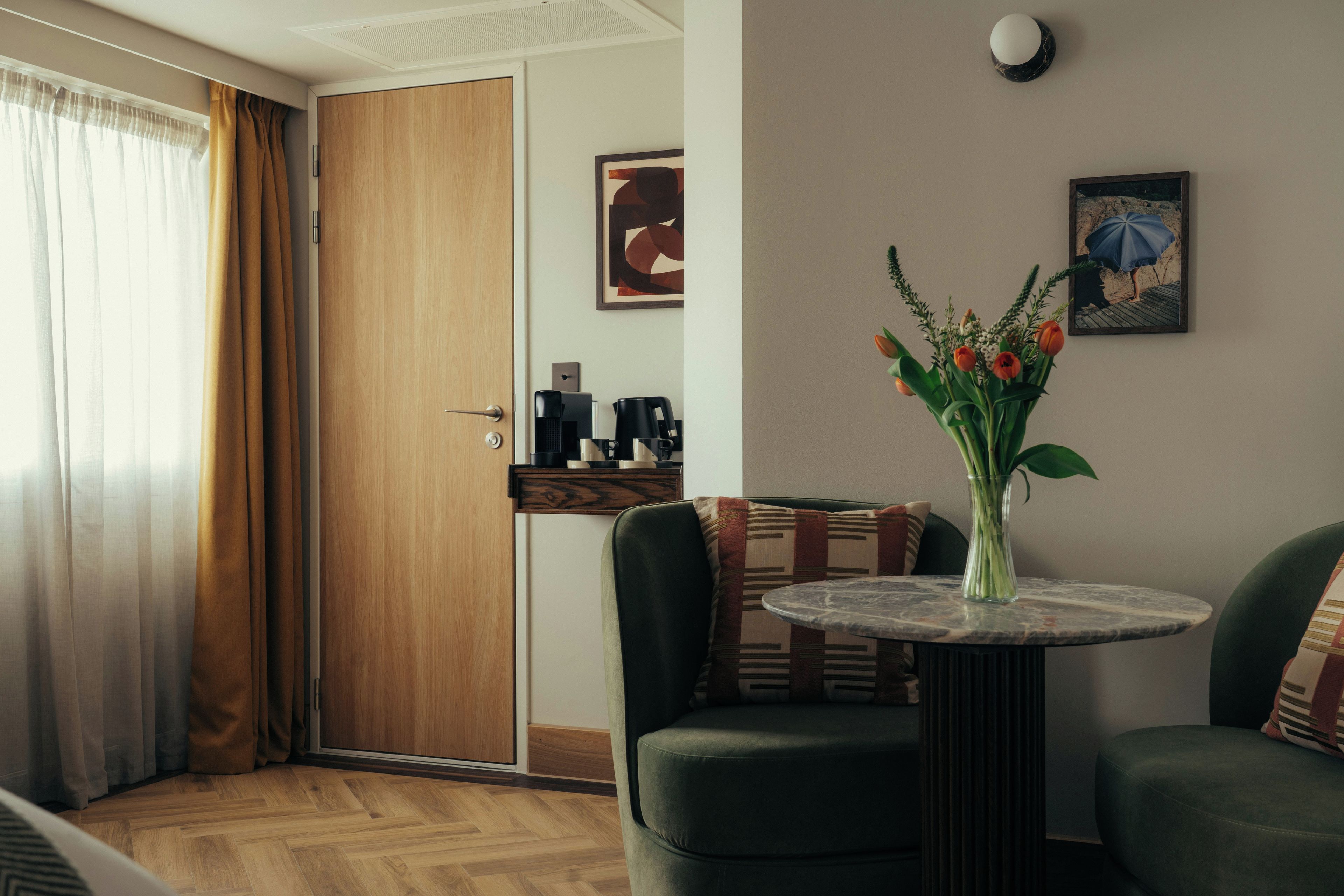 A room with a wooden door, a coffee machine, a vase with flowers, a painting, and a small table.