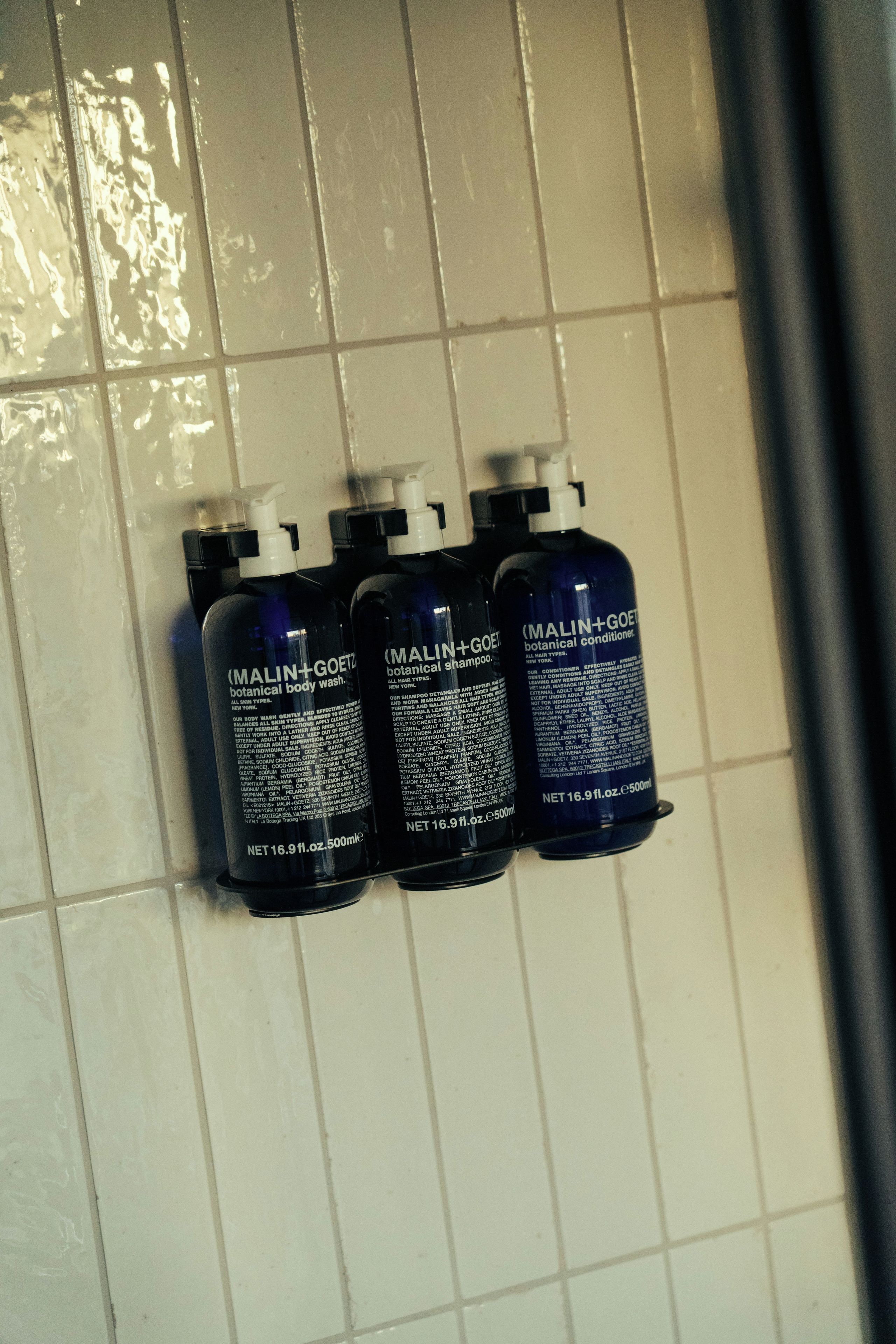 Trois bouteilles de shampoing et d’après-shampoing aux ingrédients naturels de la marque Malin+Goetz, posées sur un mur carrelé.