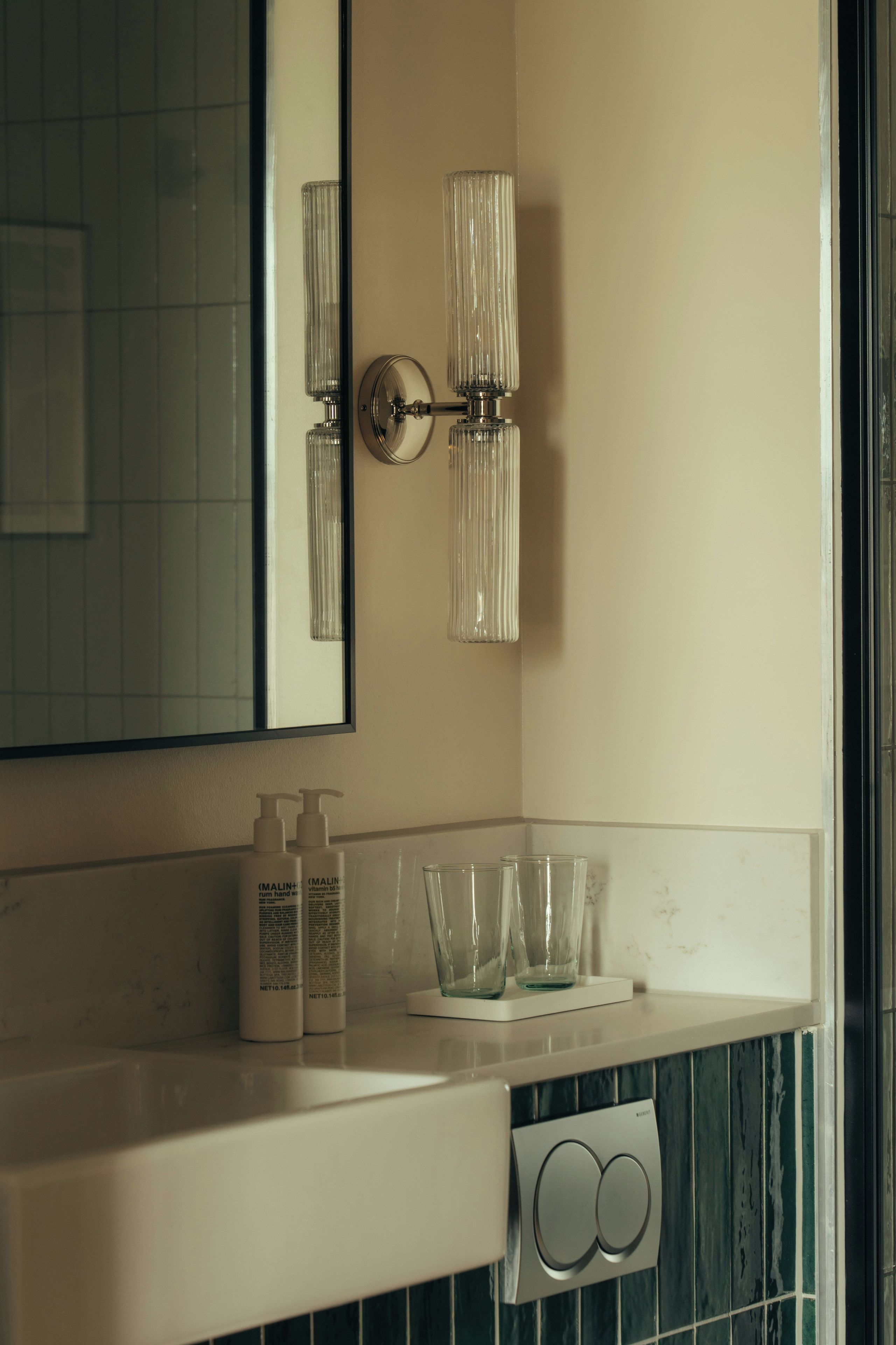 A bathroom with a mirror, two wall-mounted light fixtures, a sink with a white countertop, and a glass soap dish.