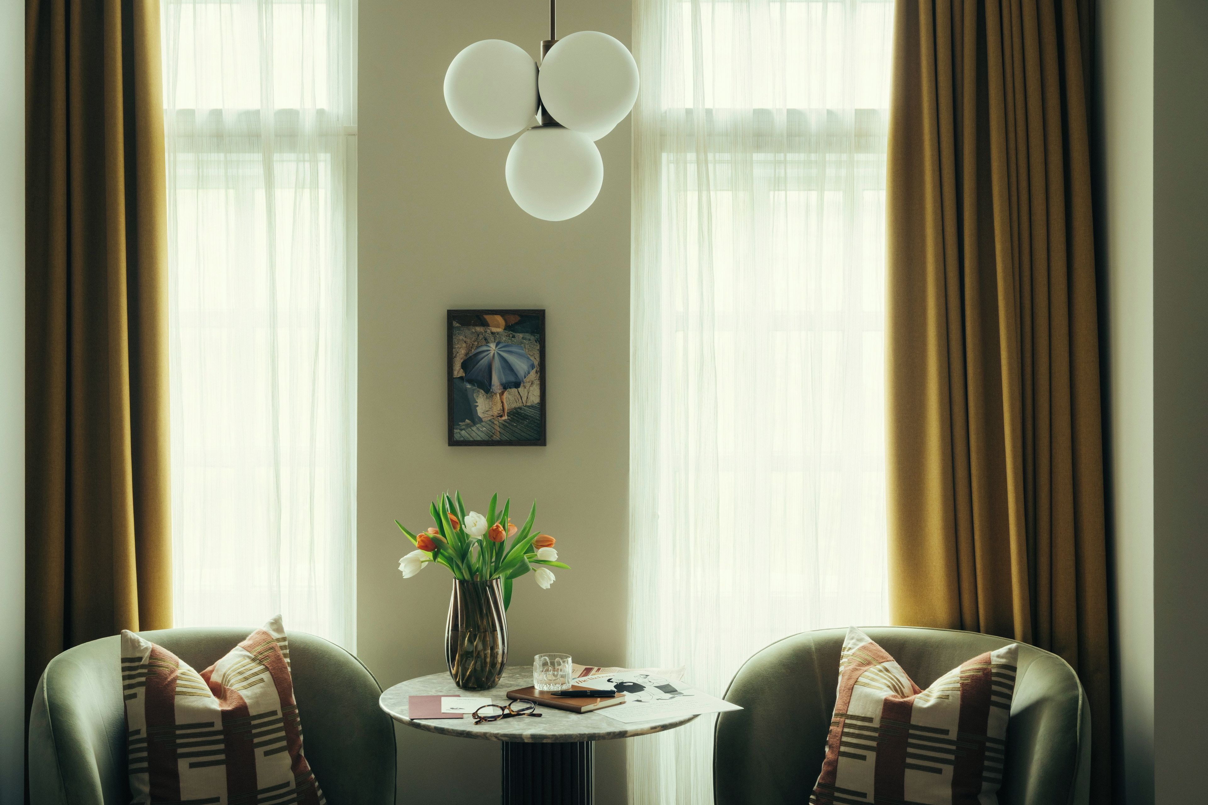Una habitación con una mesa redonda, dos sillas, un jarrón con flores, un cuadro enmarco y una lámpara colgante.