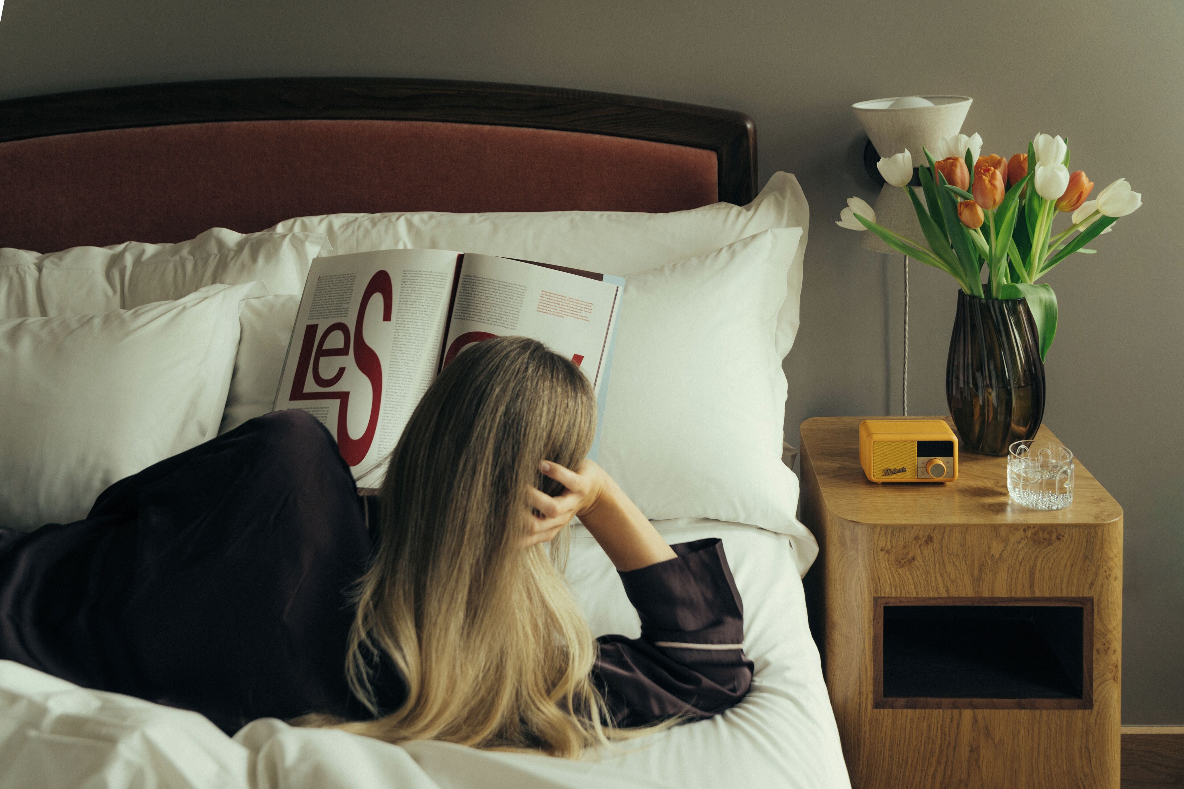 Una persona acostada en una cama con un libro, un florero con flores y un radio en la mesita de noche.