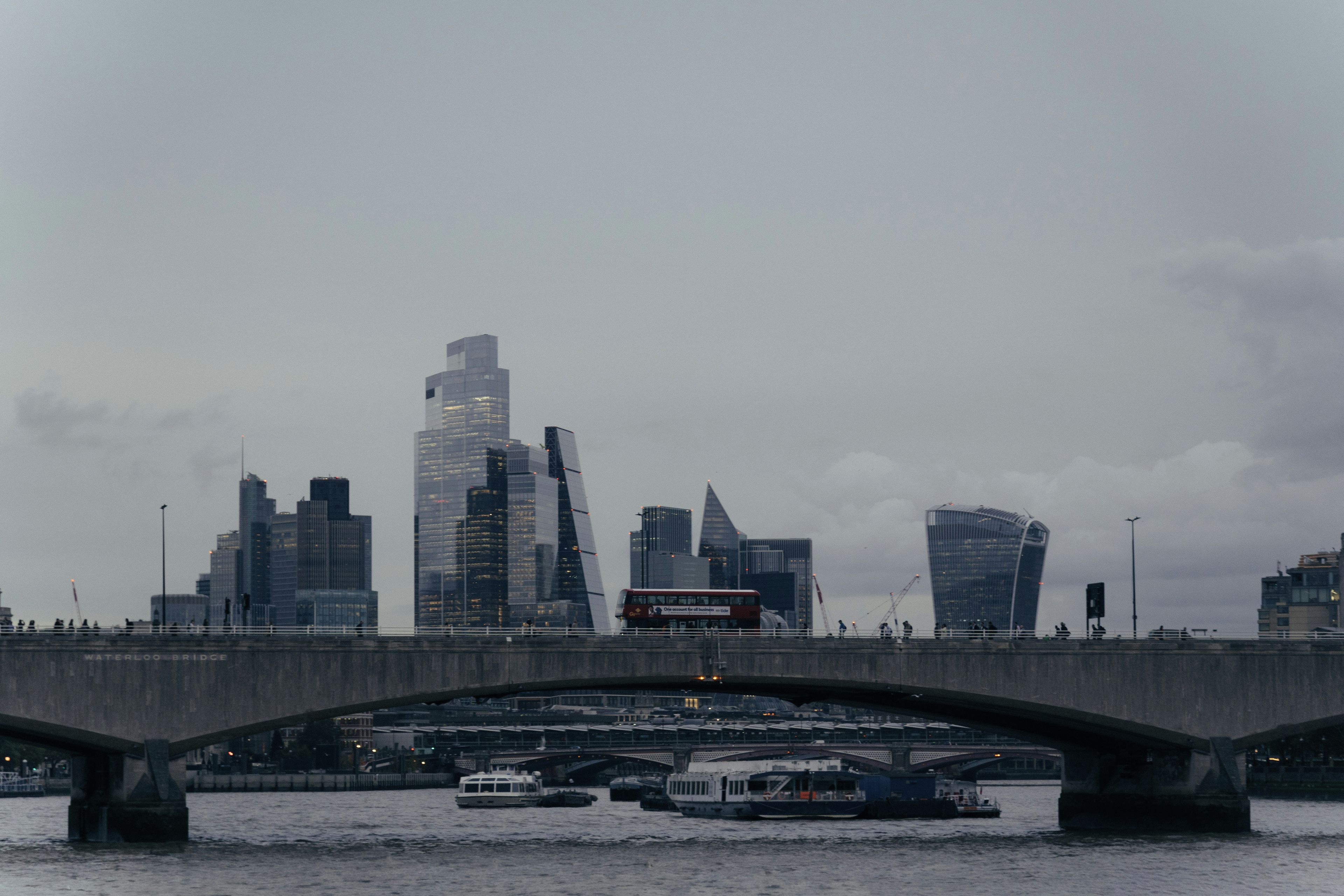 A bridge over a river with boats, a red double-decker bus, and modern buildings in the background.