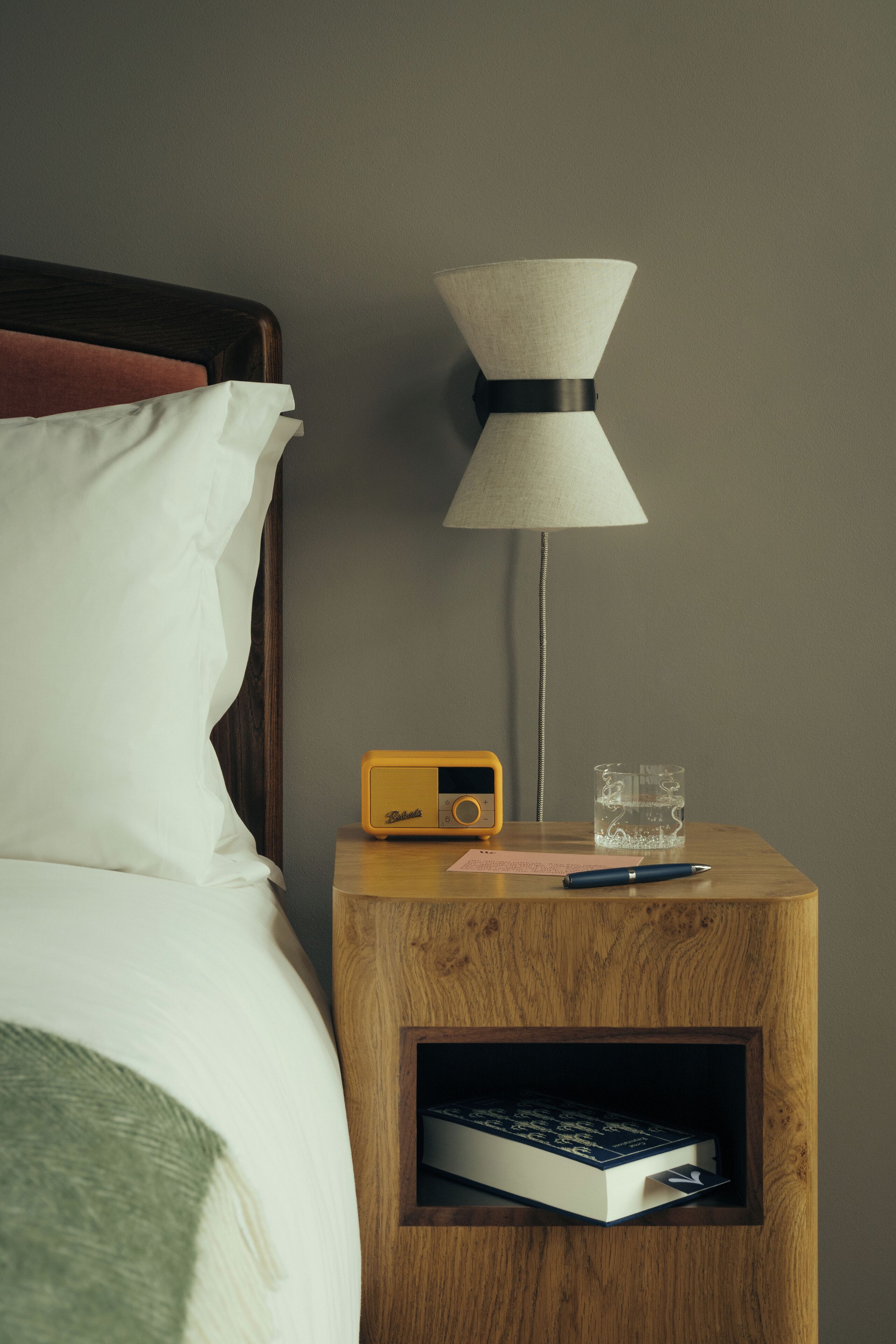 A wooden bedside table with a lamp, a radio, a glass of water, and a book.
