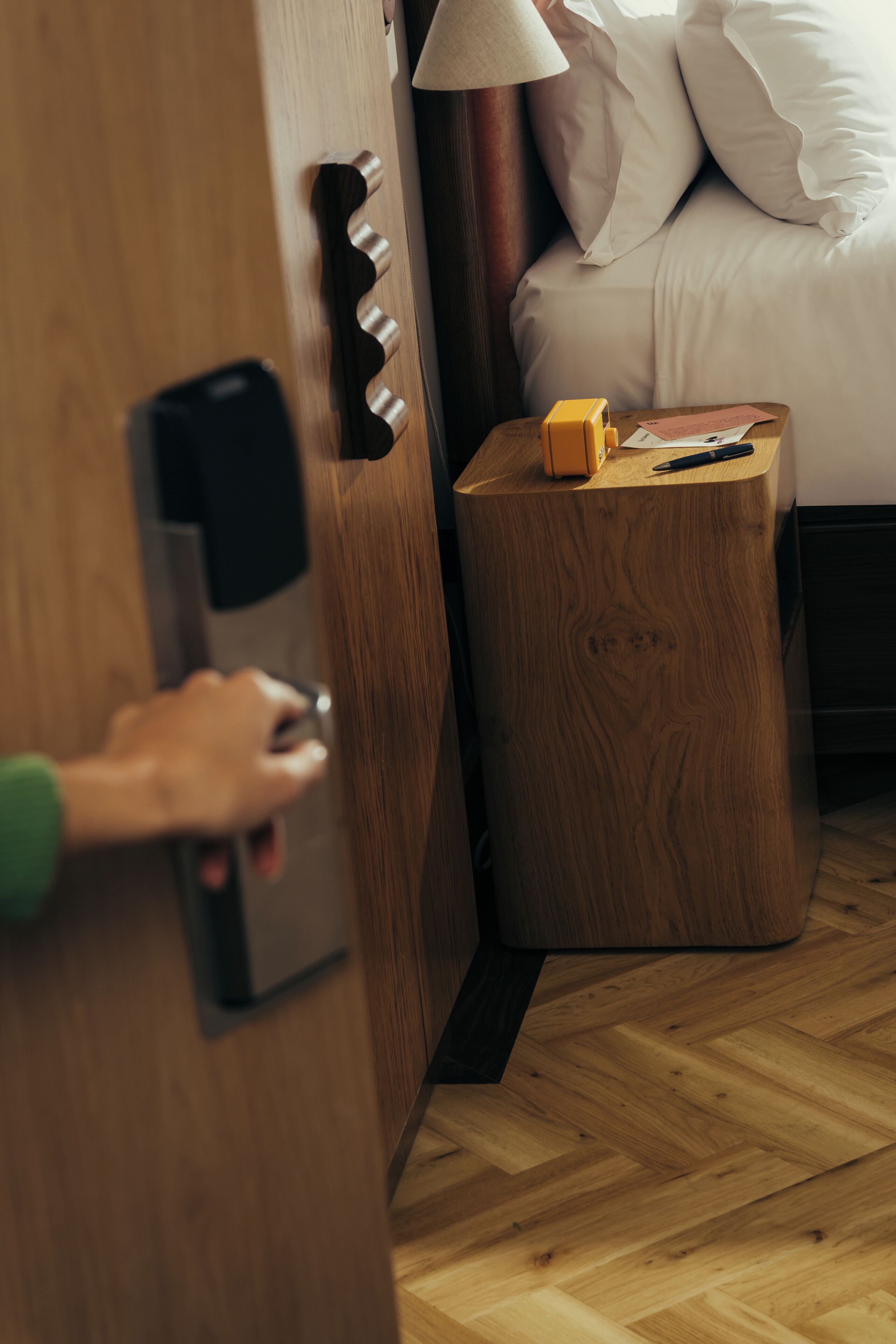 Una mano abriendo una puerta, una mesita de noche con una lámpara y una cama con almohadas.