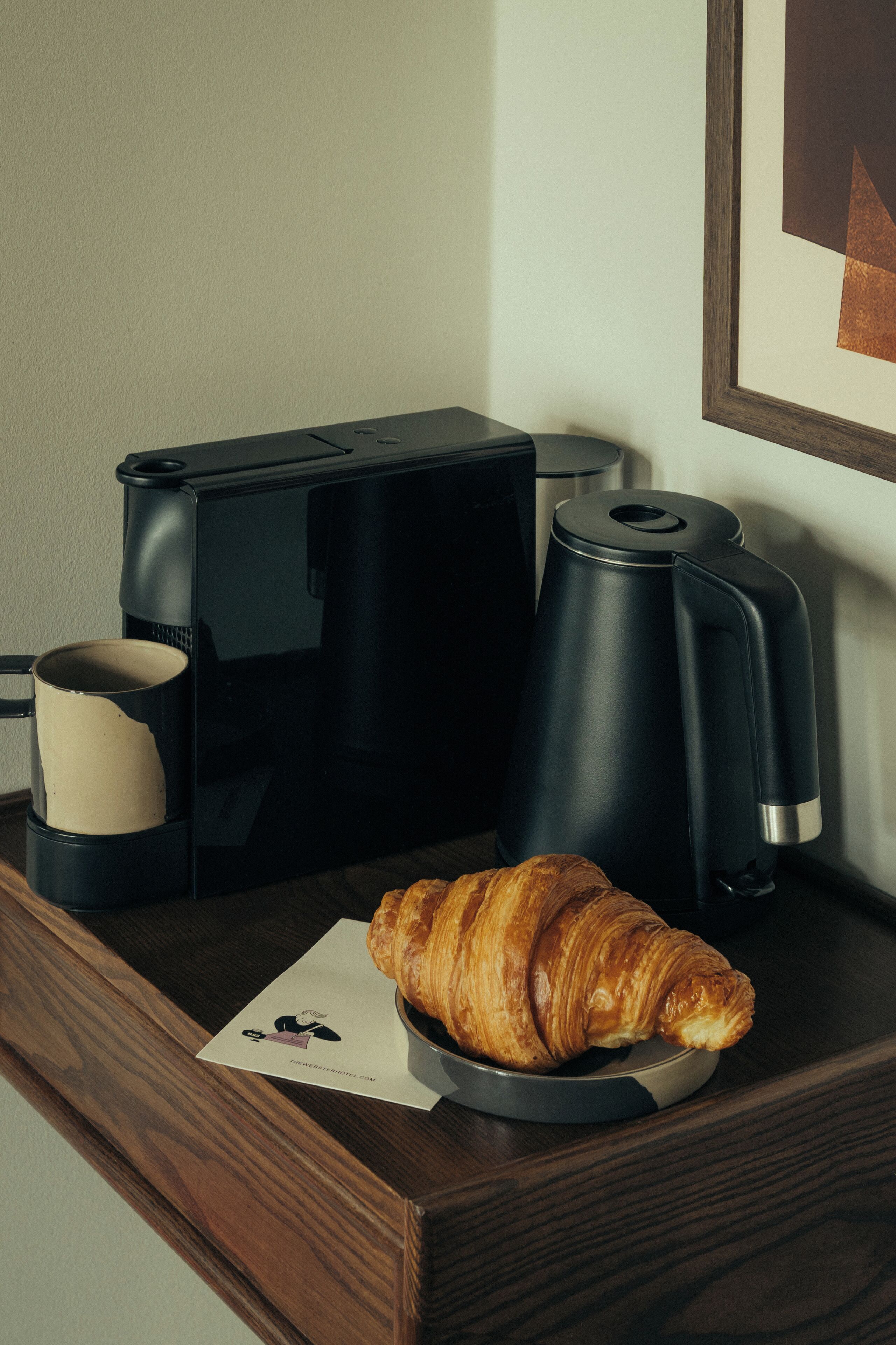 A coffee maker, a croissant, and a framed picture on a wooden surface.