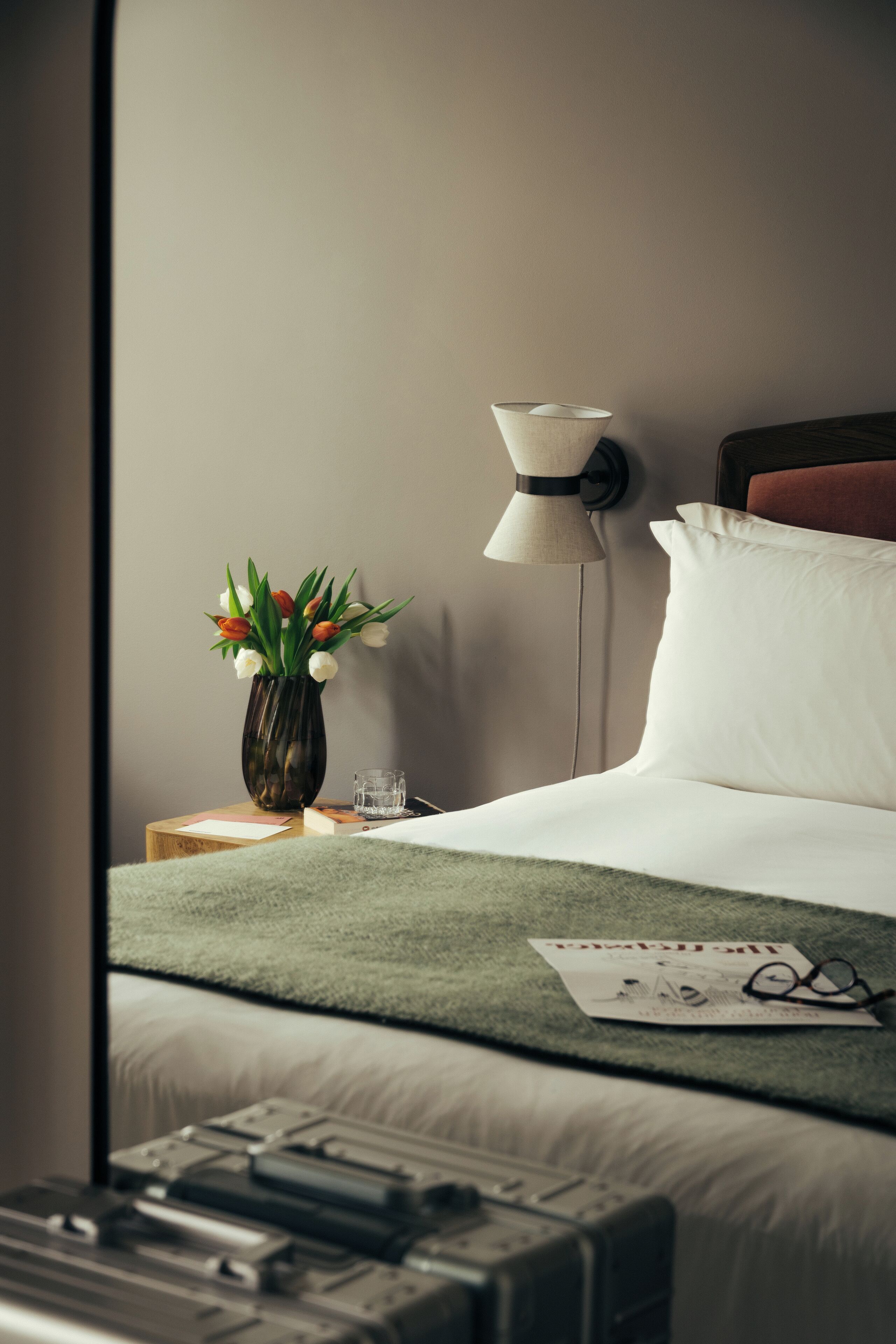 A hotel room with a bed, a bedside table with a vase of flowers, a lamp, and a window with a view.