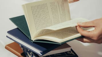 A person holding an open book with a stack of books underneath.