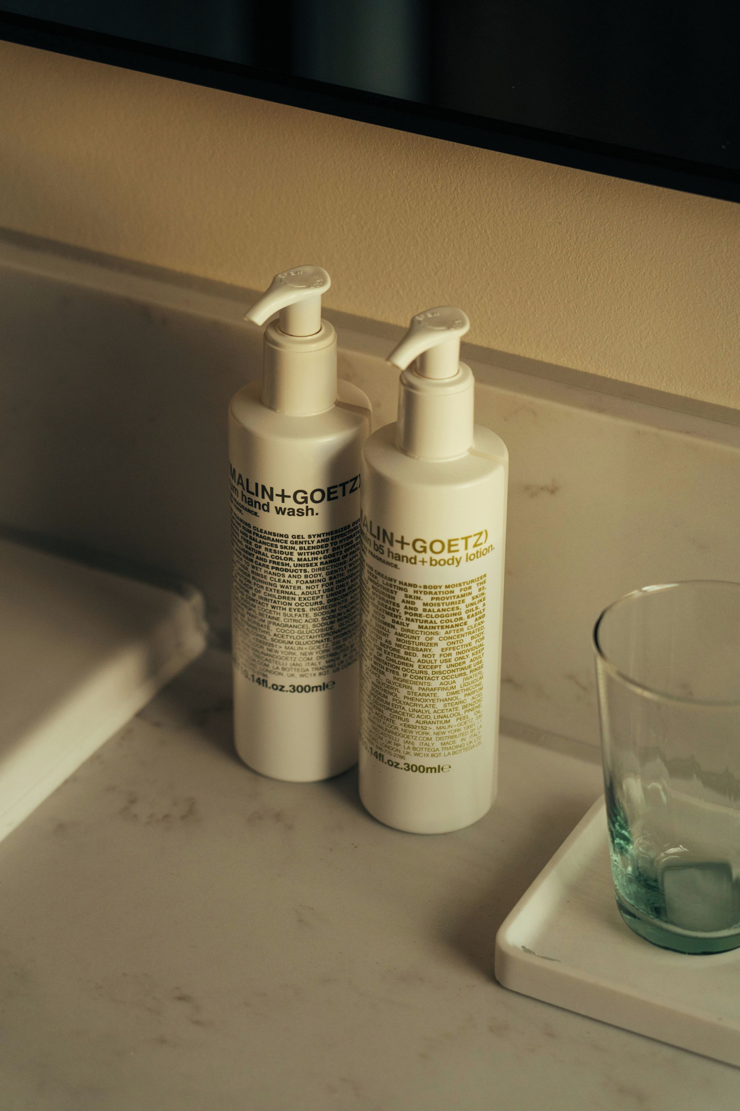 Deux flacons de lotion pour les mains et le corps de la marque MALIN+GOETZ, posés sur une surface blanche, avec un verre d’eau à proximité.