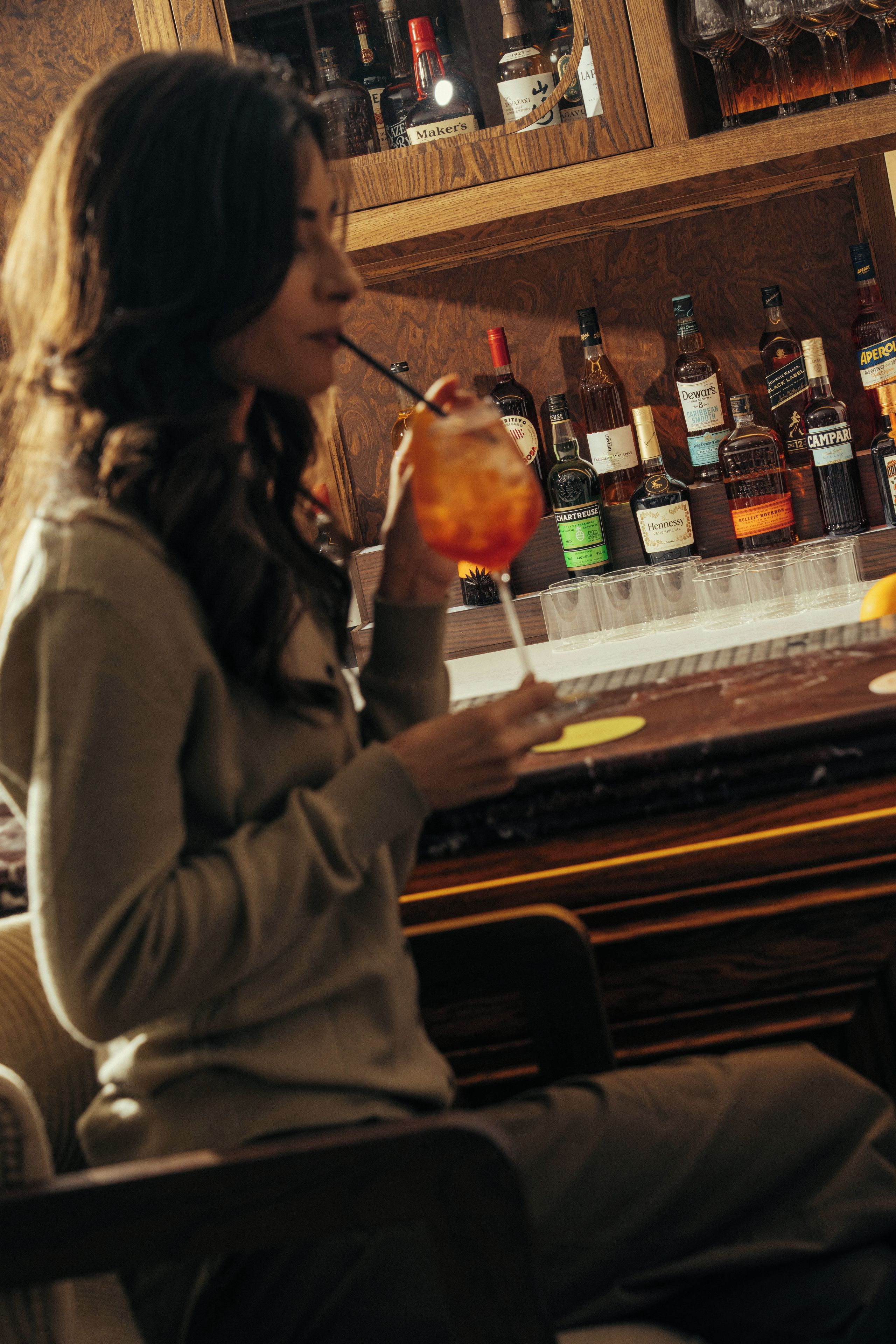 Eine Person sitzt an der Bar mit einem Getränk in der Hand, umgeben von verschiedenen Spirituosenflaschen.