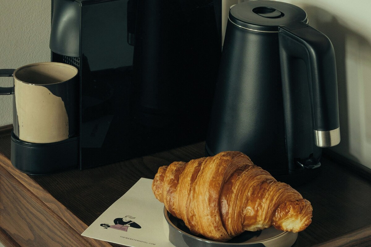 A coffee maker, a croissant, and a framed picture on a wooden surface.