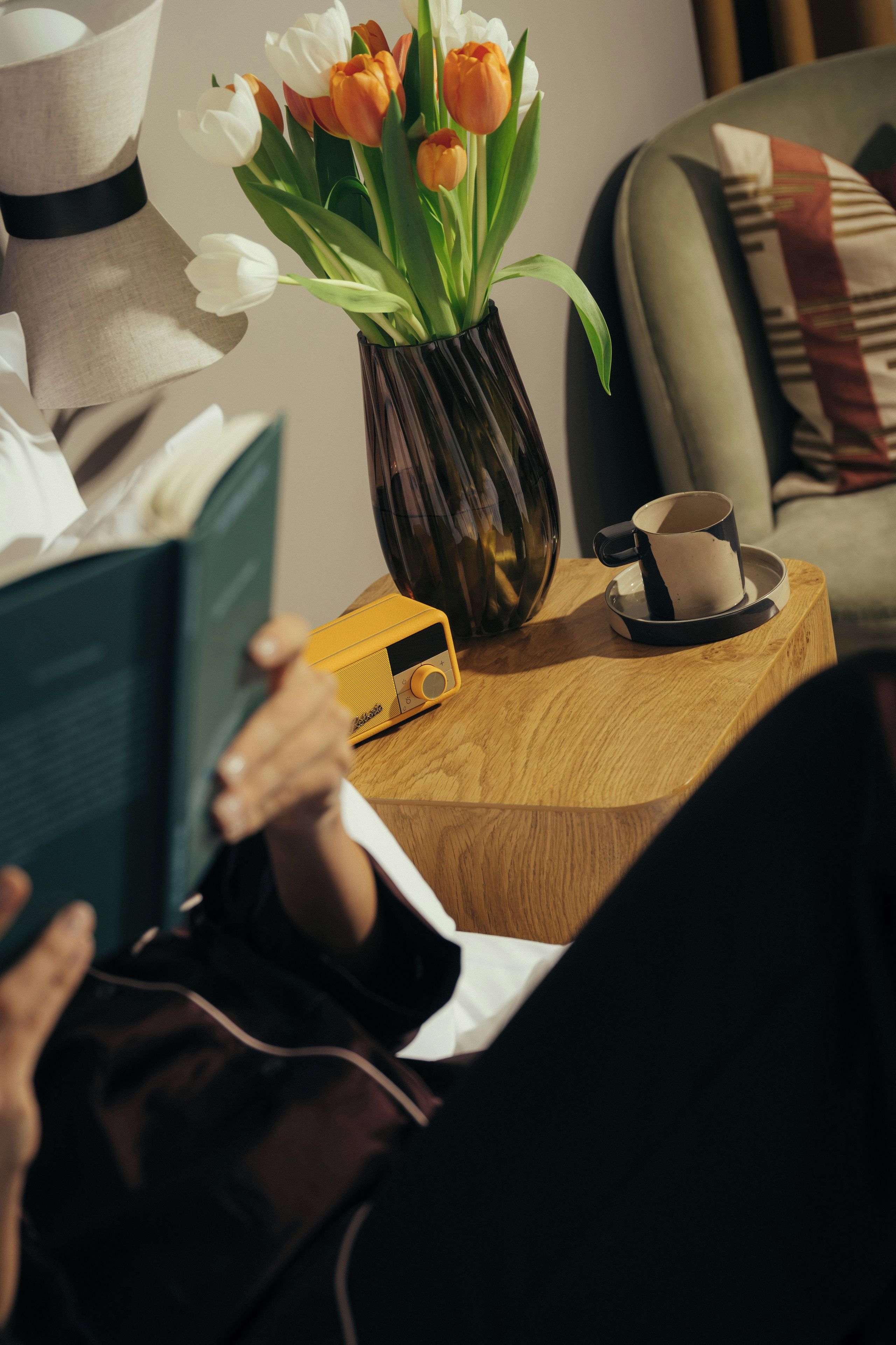 A person reading a book with a cup and saucer on a wooden side table.