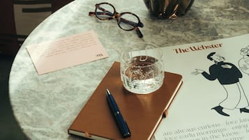 Une table ronde en marbre, sur laquelle se trouvent un carnet, un stylo, des lunettes, un verre d’eau, un vase de tulipes et un livre intitulé « The Webster ».