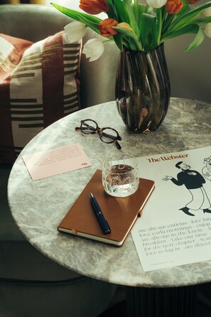 A round marble table with a notebook, pen, glasses, a glass of water, a vase with tulips, and a book titled 'The Webster'. - The Webster Covent Garden – New Lifestyle Hotel – £60 Food & Drink Credit per Stay (London)