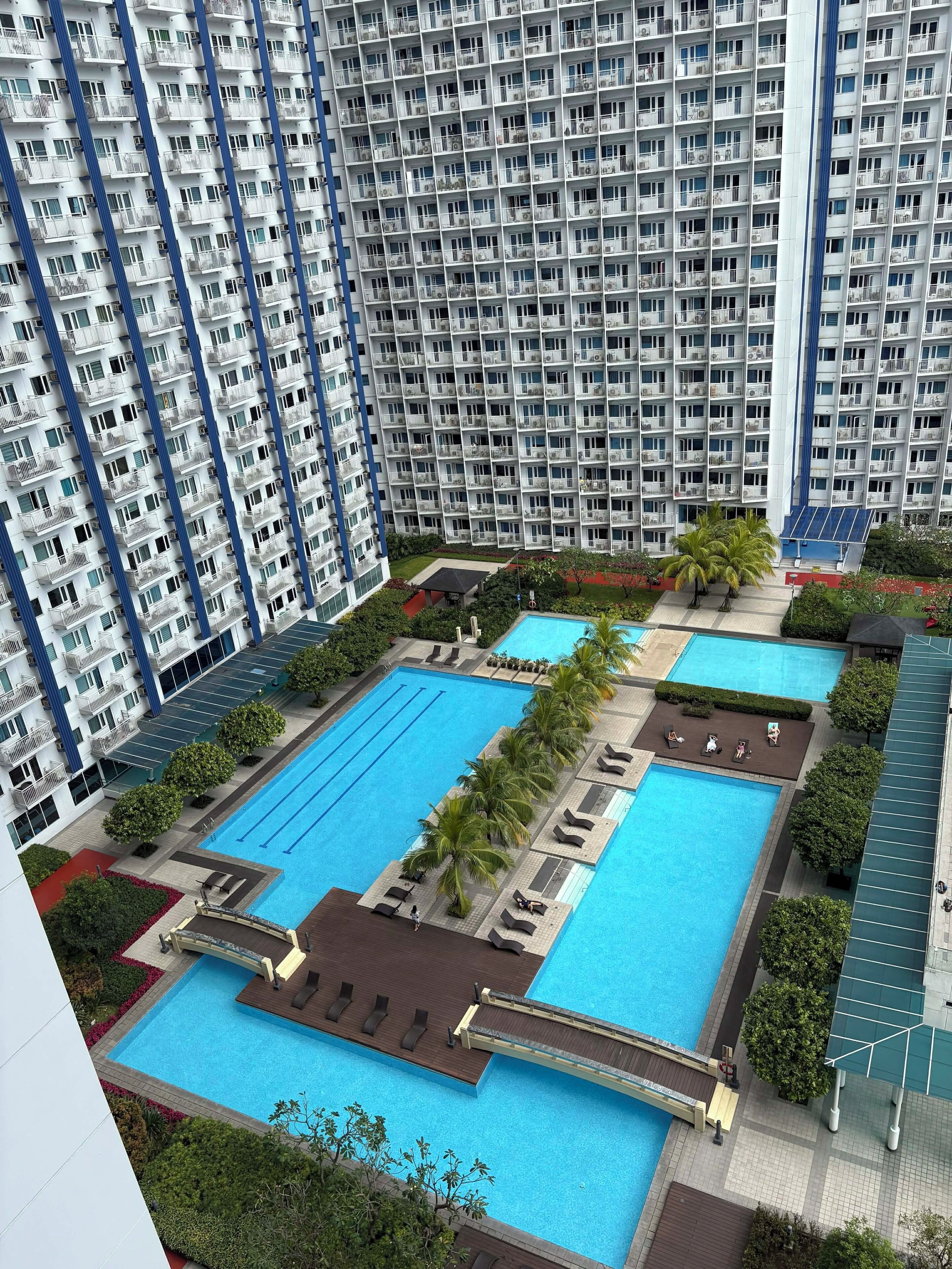 A high-rise building with multiple balconies and a large swimming pool area with sun loungers and palm trees.