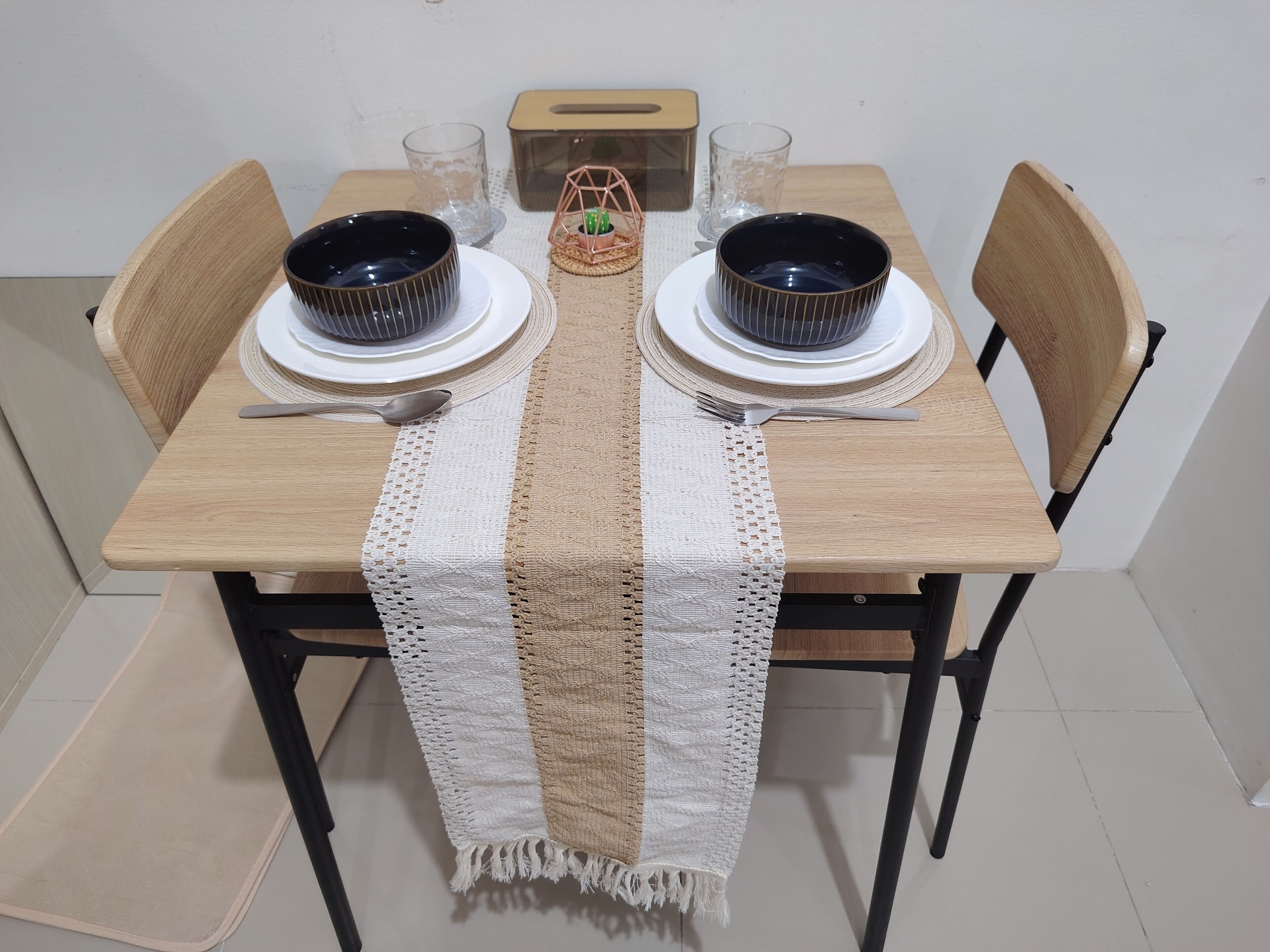 A dining table set with two black bowls, white plates, and a woven table runner.