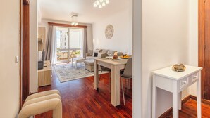 A modern living room with a dining area, wooden flooring, and a view of the city through large windows. - BoLina 2 bedroom apartment located in the historic center of Caniço. (Caniço)