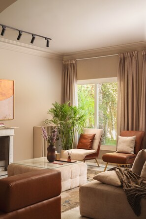 Ein Wohnzimmer mit Kamin, einem großen Fenster mit Vorhängen, einer Couch, einem Sessel und einer Topfpflanze.