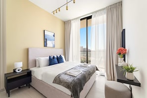 A modern hotel room with a large bed, a nightstand with a lamp, a small table with a vase of flowers, and a view of the city through the window. - Silkhaus Cozy 1bdr Binghatti Tulip JVC (Dubai)