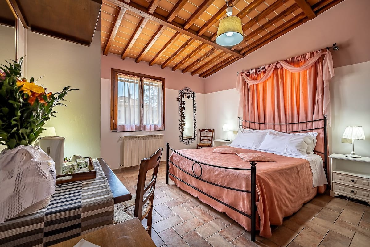 A bedroom with a bed, a chair, a table, a lamp, a mirror, and a window.