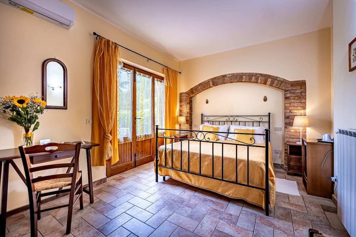 A bedroom with a bed, a desk, a chair, a window with curtains, and a stone archway.