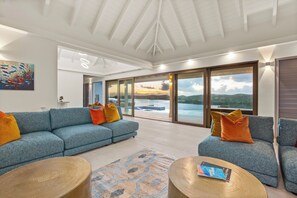A spacious living room with a blue sofa set, a round coffee table, and a large window offering a view of a body of water and hills. - Brand new luxury 5-bedroom villa on resort (FreeTown)