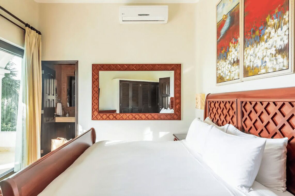 A hotel room with a bed, a mirror, a window with curtains, and a painting on the wall.