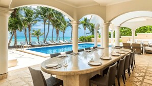 Un espace extérieur abrité où l’on peut prendre ses repas, avec une piscine et des palmiers en arrière-plan.