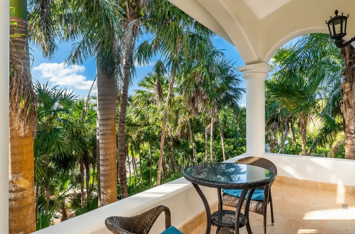 A balcony with a glass table and wicker chairs, surrounded by tall palm trees.
