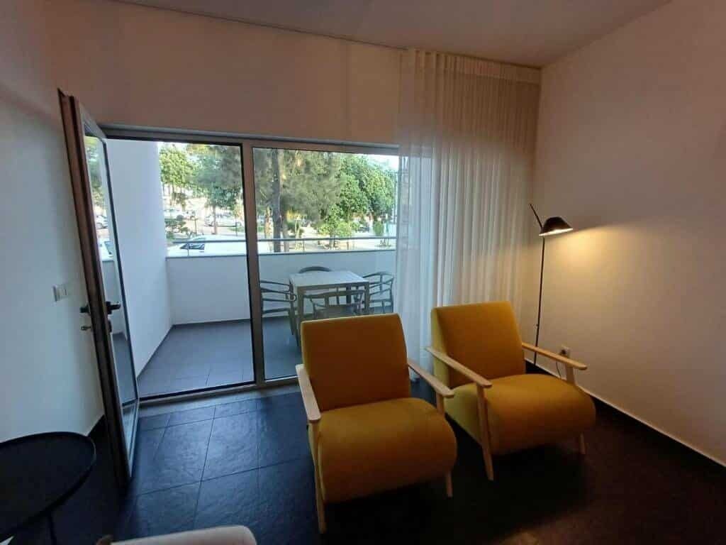 Une pièce avec deux chaises jaunes, une table, un lampadaire et une porte-fenêtre coulissante donnant sur un espace extérieur avec vue sur des arbres et un bâtiment.