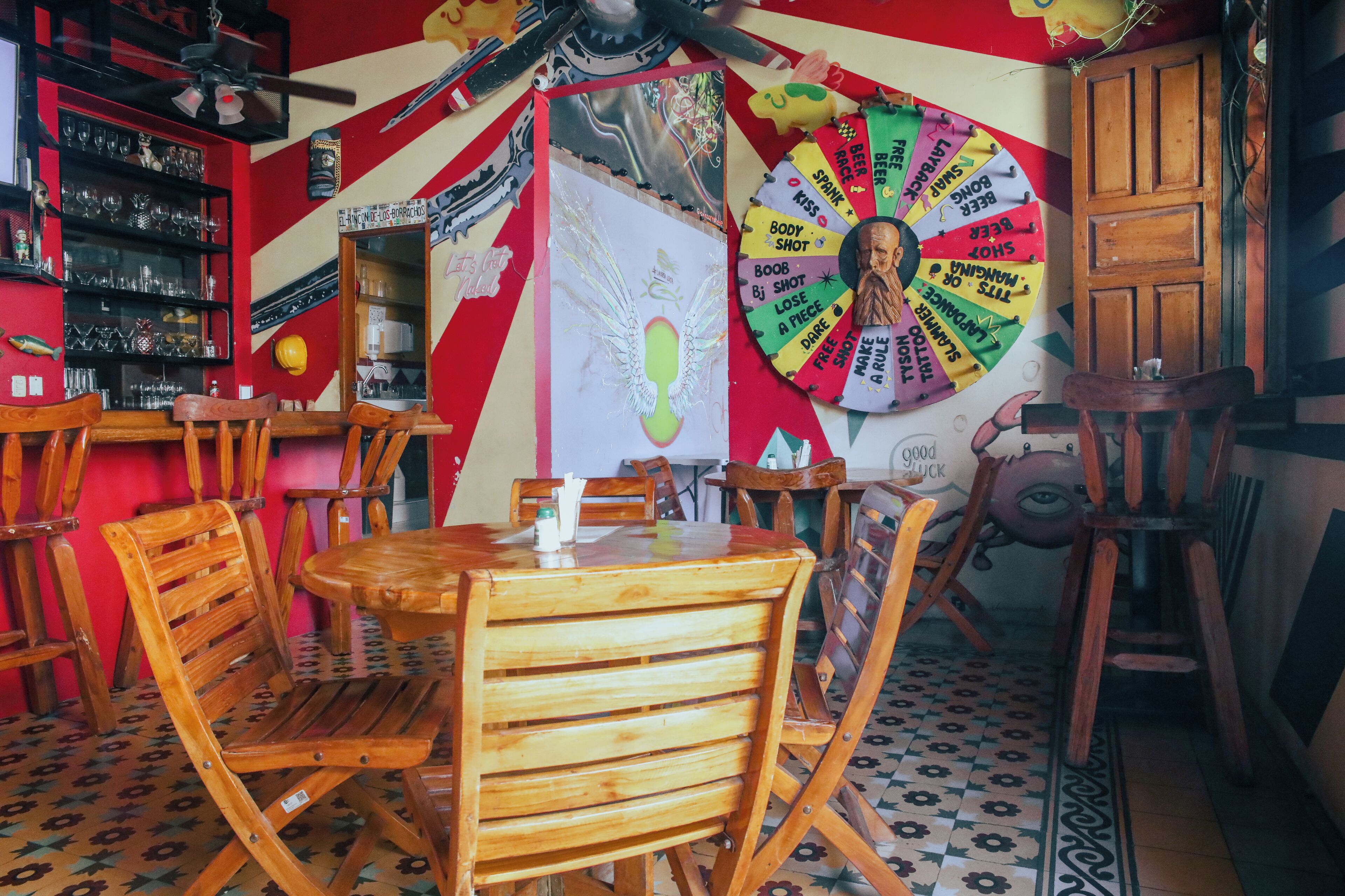 A restaurant interior with wooden tables and chairs, a colorful wall decoration, and a ceiling fan.