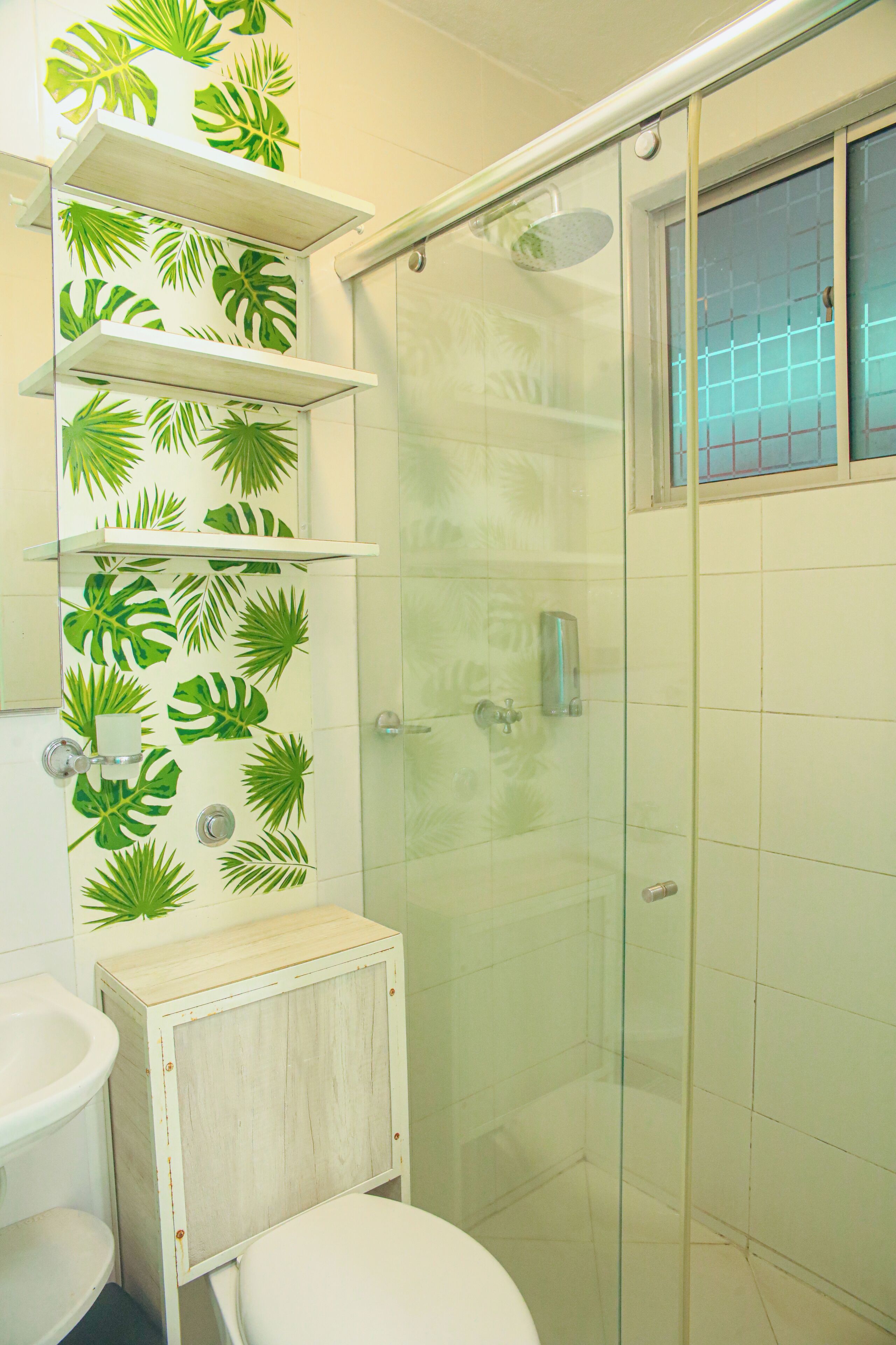Ein Badezimmer mit Toilette, Waschbecken, Dusche mit Glastür und Regalen mit Blätermotiv.