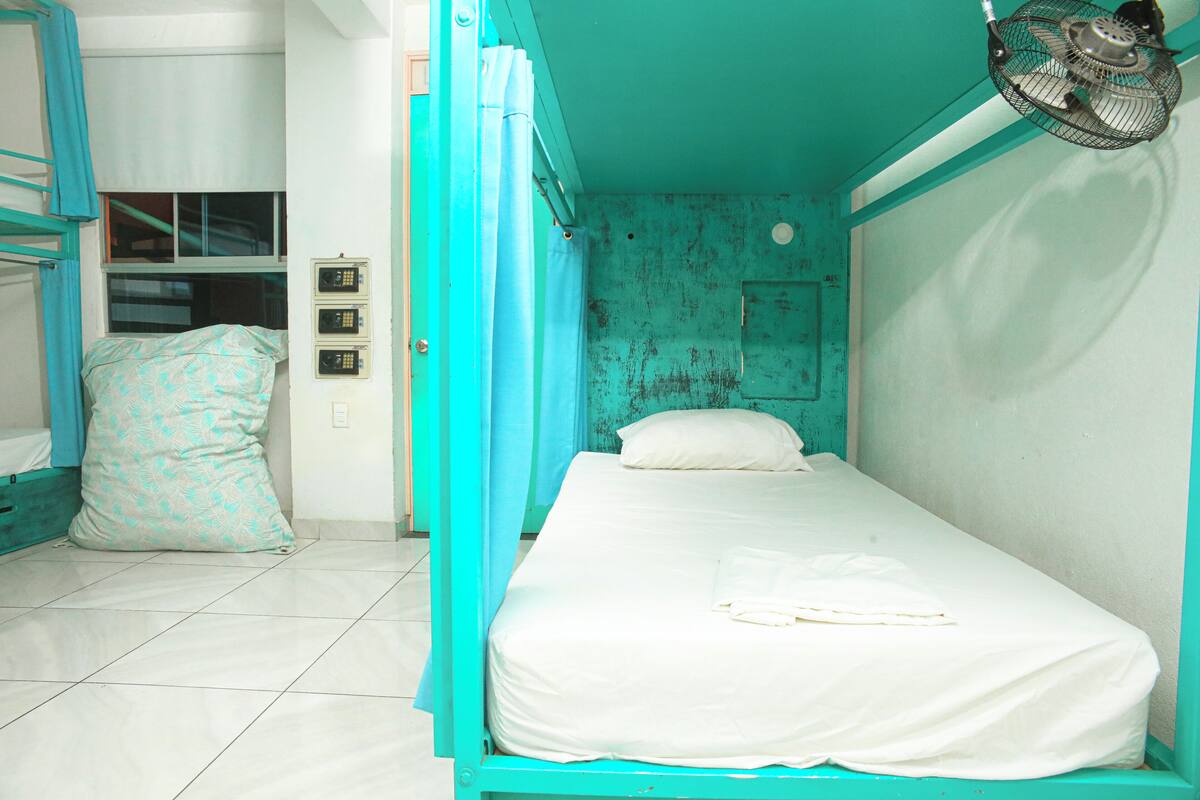 A single-bed room with a teal-colored bed frame, a white mattress, and a white pillow. There is a small window with a curtain, a wall-mounted fan, and a light switch.