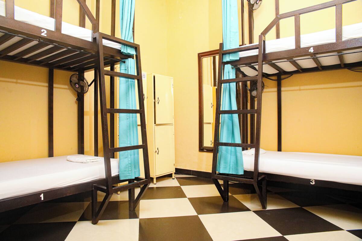 A dormitory room with bunk beds, a fan, a small cabinet, and a checkered floor.