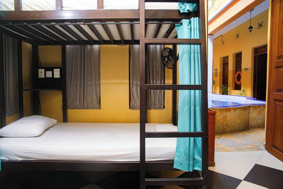 A bunk bed room with a wooden frame, a small shelf with items, and a window with curtains.