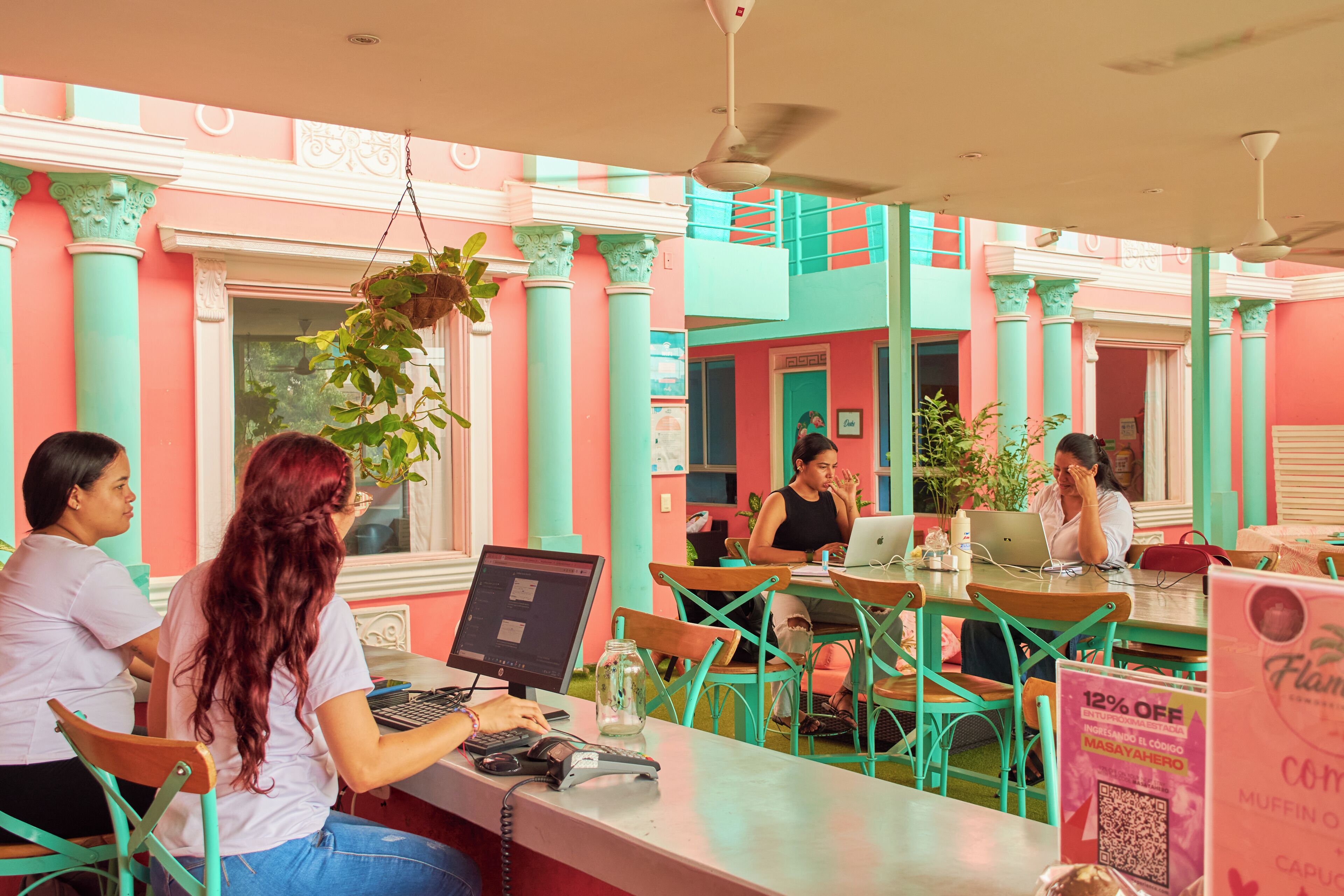 Um café com decoração em tons de verde-água e rosa, clientes usando laptops e um anúncio promocional de desconto.