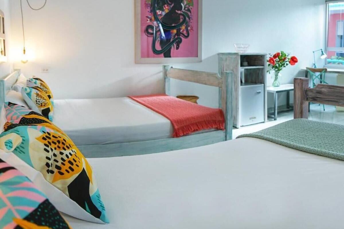 A room with two beds, a desk, a chair, a vase with flowers, and a painting on the wall.