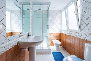 Un baño con ducha de mampara de vidrio, lavamanos, un bidé y una ventana.