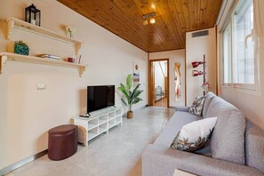 Un salon avec un téléviseur posé sur un meuble blanc, un canapé gris, un pouf marron et une plante.