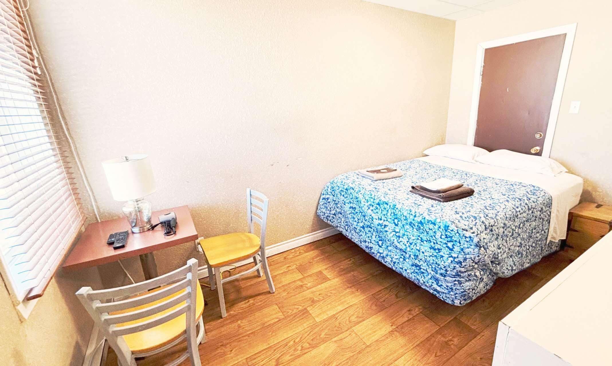 A small room with a bed, a desk, a chair, and a window with blinds.